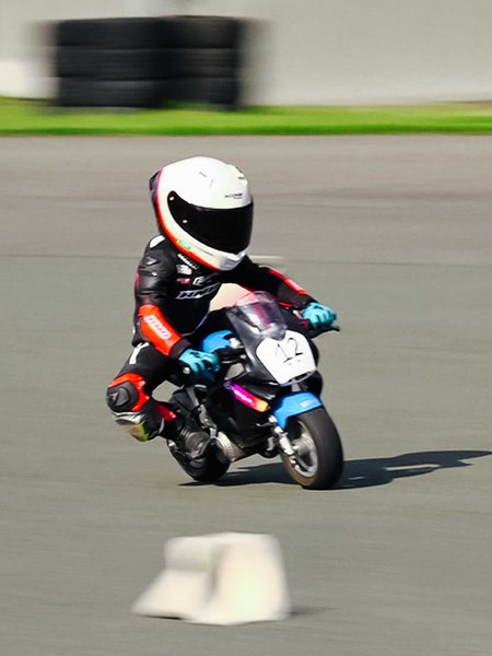 Kleiner Junge auf einem Mini-Motorrad auf einer Rennstrecke, er trägt einen weißen Helm und schwarz-rote Schutzkleidung mit blauen Handschuhen.