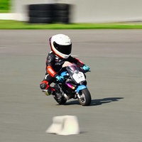 Kleiner Junge auf einem Mini-Motorrad auf einer Rennstrecke, er trägt einen weißen Helm und schwarz-rote Schutzkleidung mit blauen Handschuhen.