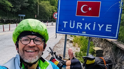 Rainer Stephan, Brille, grauer Bart und grüner Regenschutz über dem Helm, steht vor einem Landesschild der Türkei und zeigt lächelnd einen Daumen hoch in die Kamera