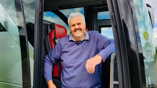 Ein Mann mit grauen Haaren und blauem Pullover steht im Eingang eines Reisebusses. Im Hintergrund rote Bussitze.