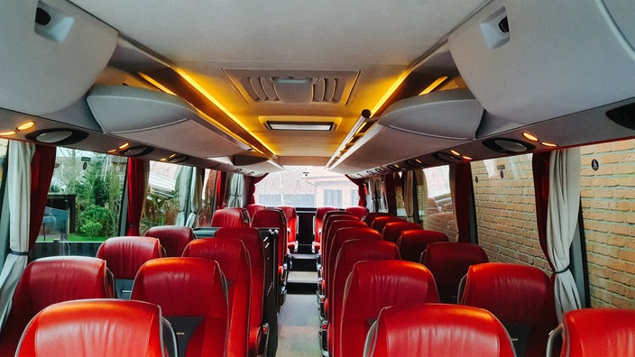 Ein Reisebus von innen. Es sind rote Sitze zu sehen und an der Decke des Busses ist indirektes gelblichen LED-Licht