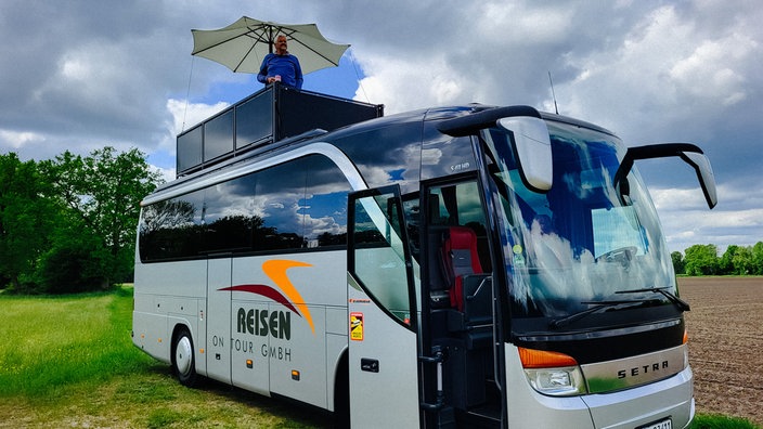 Ein XXL-Wohnmobil in Form eines Reisebusses. Auf dem Dach eine dunkle Terrasse mit einem weißen Sonnenschirm. Auf der Terrasse ein Mann mit blauem Pullover.
