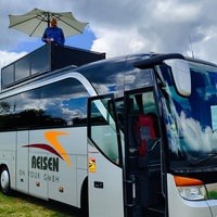 Ein XXL-Wohnmobil in Form eines Reisebusses. Auf dem Dach eine dunkle Terrasse mit einem weißen Sonnenschirm. Auf der Terrasse ein Mann mit blauem Pullover.