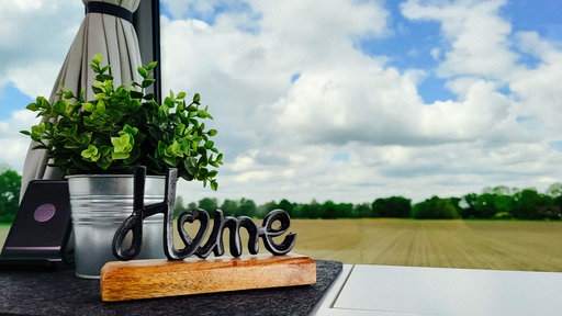 Eine hölzerne Kücheplatte. Hinter ihr ein Fenster, welches mitten in die Natur zeigt. Auf der Kücheplatte ein Dekoschild mit der Schrift "Home" 