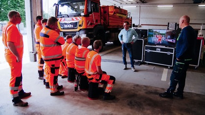 Straßenwärter in orangener Kleidung stehen in einer Halle vor einem Monitor und zwei Schulungsleitern für Arbeitssicherheit.