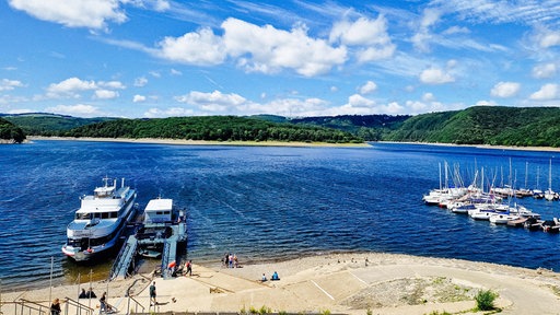 Auf dem leuchtend blauen Rursee liegt das Passagierschiff "Stella Maris" am Anleger vor dem Sandstrand.