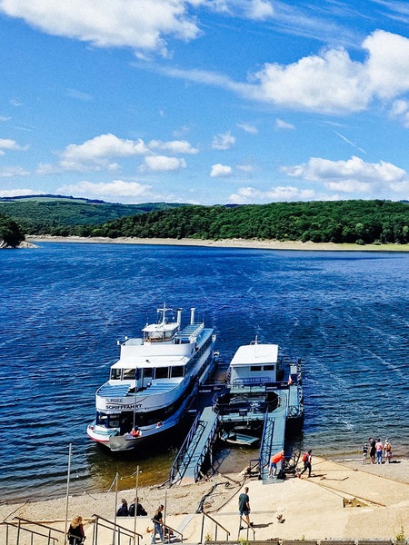 Auf dem leuchtend blauen Rursee liegt das Passagierschiff "Stella Maris" am Anleger vor dem Sandstrand.