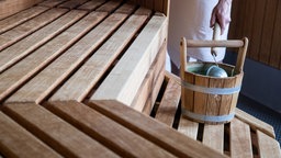 Mensch steht in einer Holzsaune hält Holzkelle und schöpft Wasser aus Holzeimer