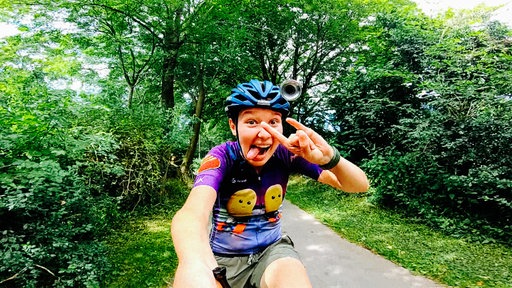 Eine junge Frau mit Helm fährt schnell auf einem Longboard, sie lacht in die Kamera und streckt die Zunge raus