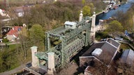 Man sieht das Schiffshebwerk in Henrichenburg über einem Fluss, mit vier Türmen an den Ecken. Am Fluss entlang sieht man Bäume und Häuser.