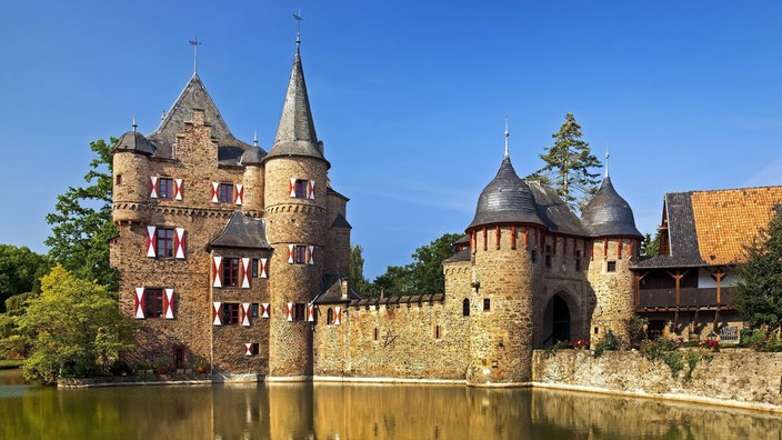 An einem Teich gelegene Burg aus braunem Stein mit grauem Dach. Ein Torhaus mit zwei Türmen.