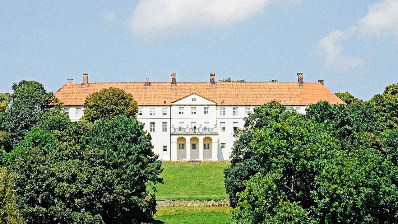 Schloss Cappenberg mit rotem Dach und weißer Fassade. Im Hintergrund Wald und Felder.