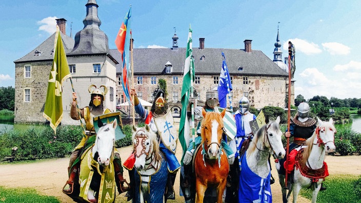 Fünf Ritter auf ihren Pferden stehen mit erhobenen Fahnen vor Schloss Lembeck.