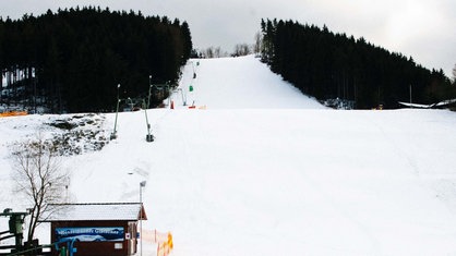 Eine breite, ruhige Skipiste, die von dichten Waldstreifen eingerahmt wird, mit einem einfachen Skilift und einer kleinen Holzhütte im Vordergrund.