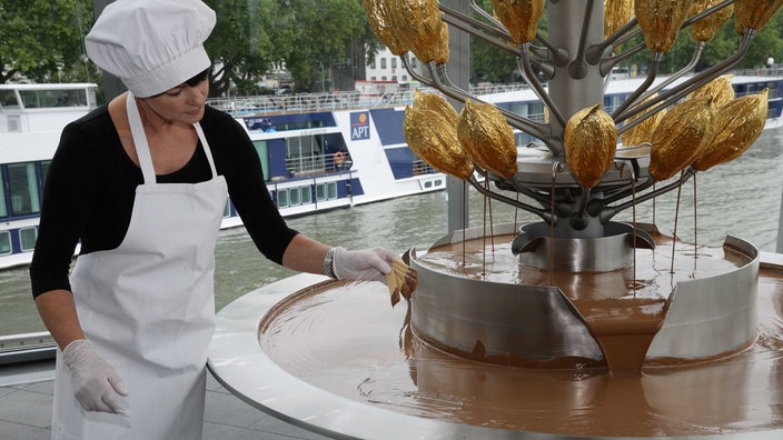 Eine Frau mit weißem Kochhut und weißer Schürze steht an einem Brunnen, in dem Schokolade runterläuft, sie hält vier Waffeln in der Hand, die sie gerade in die Schokolade getaucht hat