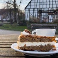 Ein Stück Kuchen auf einem Teller in Nahaufnahme. Im Hintergrund der Bauernhof Schultenhof.
