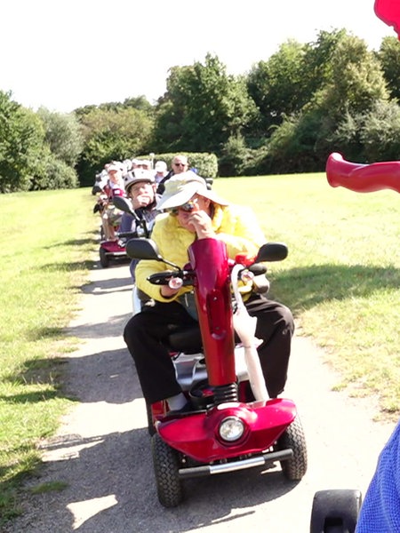 Wolfgang Schwan sitzt in einem Park auf seinem Elektroscooter, im Hintergrund weitere Mitglieder der Duisburger Scootergang.