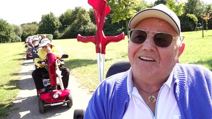 Wolfgang Schwan sitzt in einem Park auf seinem Elektroscooter, im Hintergrund weitere Mitglieder der Duisburger Scootergang.