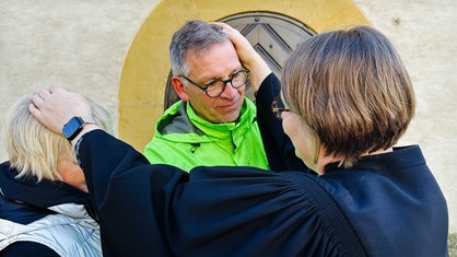 Kerstin Pilz, kurze Haaren und schwarze Robe, hält jeweils eine Hand auf die Köpfe von Rainer und Marion Stephan 
