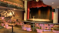 Blick auf eine kleine Theaterbühne, auf der nur ein kleiner Stehtisch steht. Rote Vorhänge zieren die Bühne. Vor ihr stehen rosafarbene, leere Stühle.