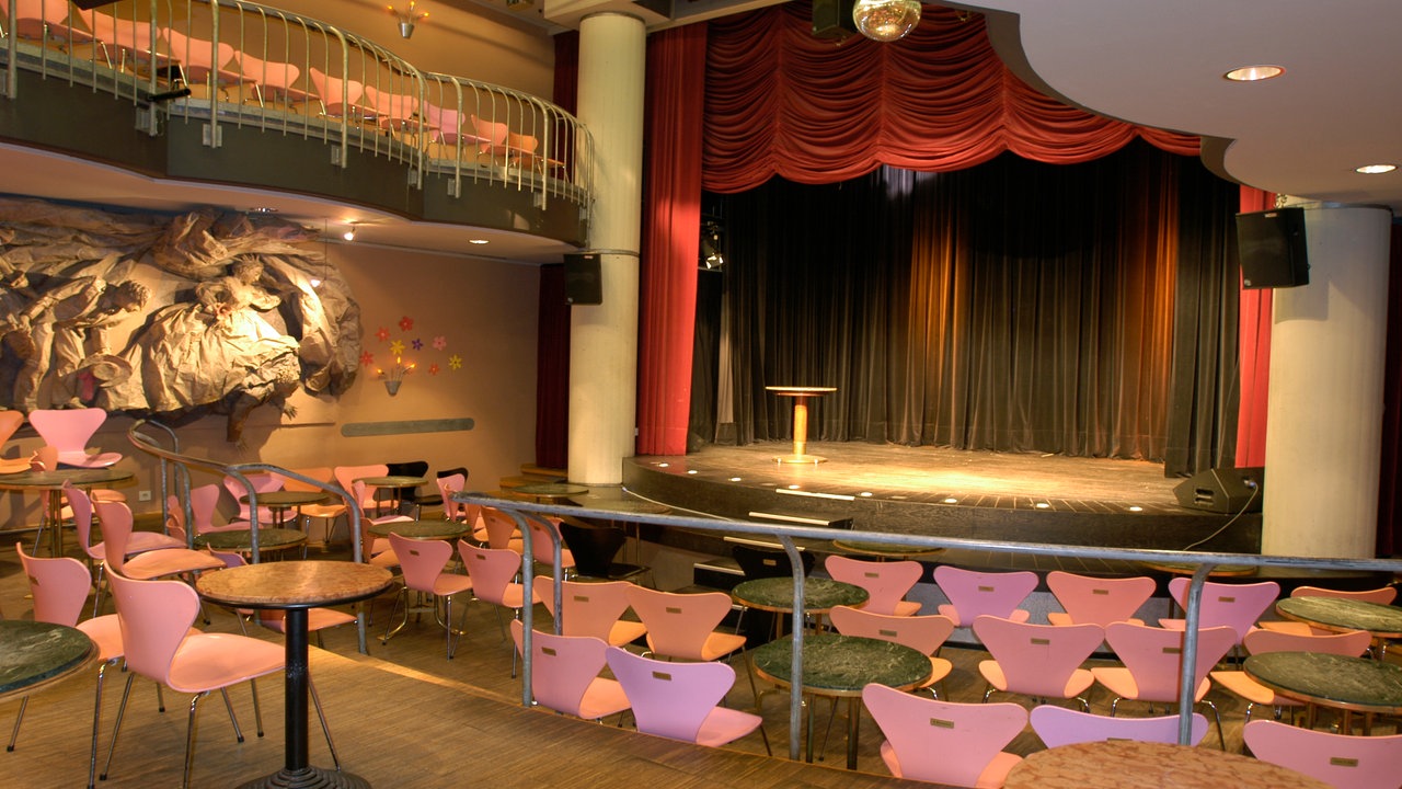 Blick auf eine kleine Theaterbühne, auf der nur ein kleiner Stehtisch steht. Rote Vorhänge zieren die Bühne. Vor ihr stehen rosafarbene, leere Stühle.