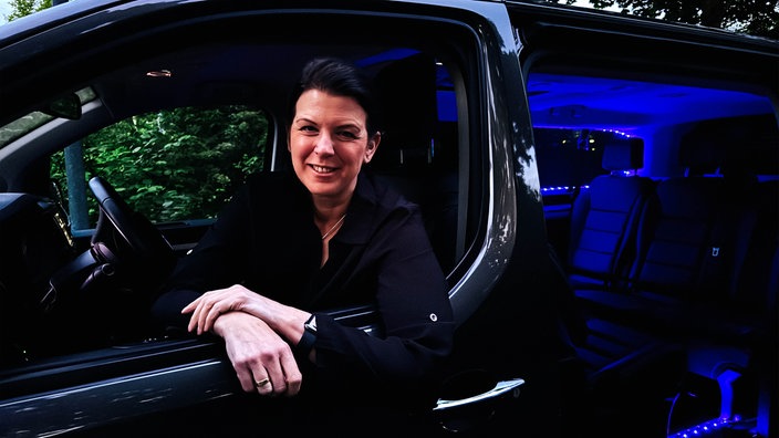 Kerstin Schulz-Höfel sitzt in ihrem schwarzen Bulli, den sie zu einem Party-Shuttle umfunktioniert hat. Im Inneren des Autos sind blaue LED- Lichter.