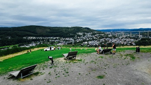 Ein paar Menschen befinden sich auf einem geschotterten Platz auf einem Hügel, im Hintergrund sieht man ins Tal hinab