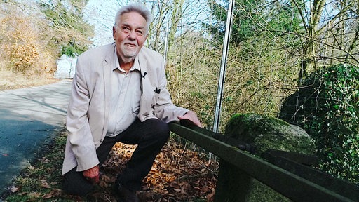 Werner Zahn, ein grauhaariger Mann mit Bart im weißen Hemd und hellen, karierten Sakko, hockt links neben einer alten Straßenbahnschiene im Wald.