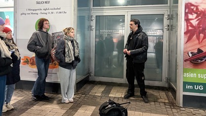 Markus R., in Schwarz gekleidet, steht vor dem überfachten Notausgang von Galeria Kaufhof, links neben ihm stehen Teilnehmende der Stadtführung, denen er zugewandt ist