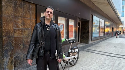 Markus R., komplett in Schwarz gekleidet, steht vor den Schaufenstern von Galeria Kaufhof in der Kölner Innenstadt, er trägt eine schwarze Tasche um die Schulter und einen schwarzen Gehstock, rechts hinter ihm steht ein Rad