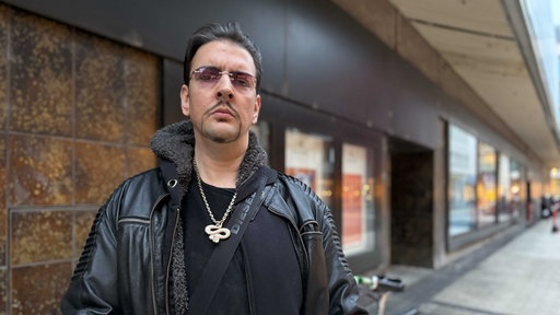 Markus R., komplett in Schwarz gekleidet, einen schwarzen Taschengurt um die Schulter, goldene Schlangen-Kette um den Hals, rosa-gefärbte Brille.