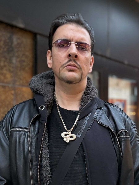 Markus R., komplett in Schwarz gekleidet, einen schwarzen Taschengurt um die Schulter, goldene Schlangen-Kette um den Hals, rosa-gefärbte Brille.