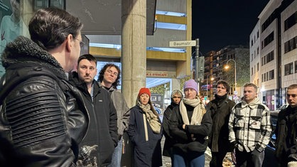 Markus R. steht einer Gruppe von acht Menschen zugewandt, die im Halbkreis um ihn herum stehen und ihn anschauen, im Hintergrund das Straßenschild "Baumstr."