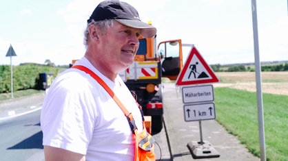 Ein Mann in Arbeitskleidung steht hinter seinem Unimog auf der Straße.
