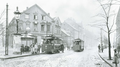 Zwei kleine Straßenbahnen fahren durch die Stadt. Alte Häuser, Menschen in Hüten und Mänteln.