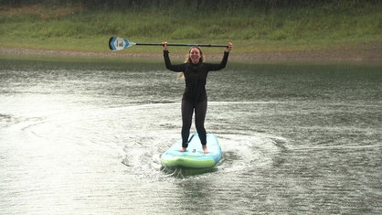 Eine junge Frau steht auf einem Stand-up-Paddling-Board auf dem Wasser und reckt ihr Paddel über ihren Kopf.