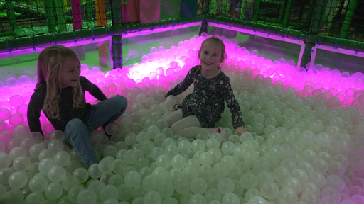 Zwei junge Mädchen spielen in einem Bällebad mit weißen Bällen. Ein pinkes Neonlicht umringt das Bällebad. 