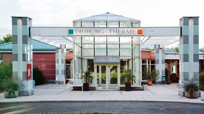 Ein recheckiger Glasbau mit dem Schriftzug "Driburg Therme". An jeder Ecke steht eine hohe Steinsäule.