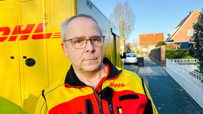 Ein Mann mit grauen Haaren und Brille, steht in DHL-Jacke vor einem großen Lieferwagen, ebenfalls von DHL.