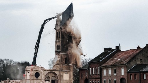 Der letzte Turm des Immerather Doms Sankt Lambertus wird abgerissen. Dabei fällt gerade die Turmspitze auf den Boden. Rechts davon leerstehende Wohnhäuser.