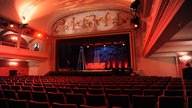 Blick auf eine rot-orange beleuchtete Theaterbühne vom Publikum aus. Der Saal ist leer, auf der Bühne selbst stehen zwei Leitern.