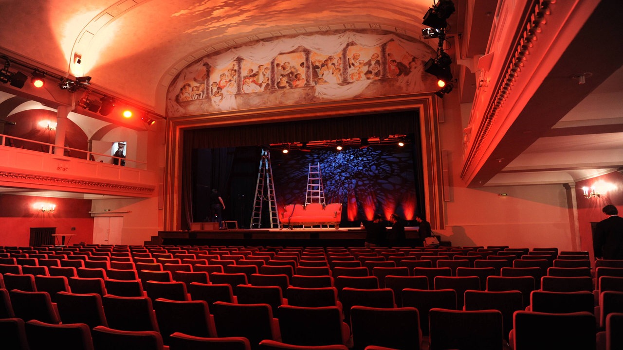 Blick auf eine rot-orange beleuchtete Theaterbühne vom Publikum aus. Der Saal ist leer, auf der Bühne selbst stehen zwei Leitern.