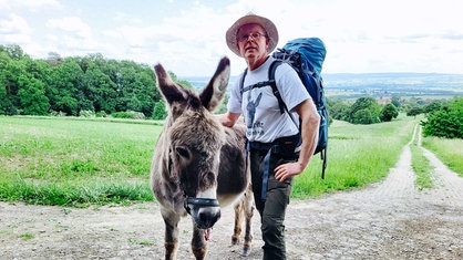 Klaus Lange steht neben seinem Esel in der Natur. Lange trägt einen Hut und einen Wanderrucksack. 
