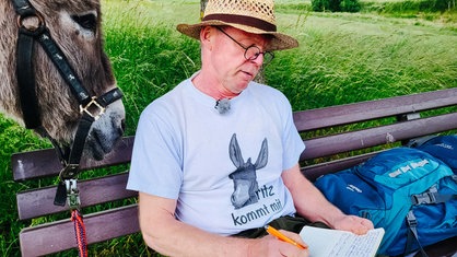 Klaus Lange sitzt auf einer Bank und macht Notizen. Er trägt ein T-Shirt mit der Aufschrift "Fritz kommt mit". Hinter ihm steht Esel Fritz.