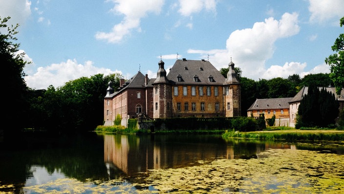 Ein Schloss in roten und gelben Farben. Es ist umgeben von Wasser. Im Hintergrund Bäume.