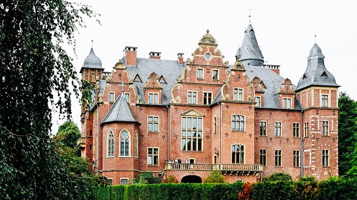 Ein Schloss mit einer rötlichen Fassaden und vielen Türmchen, die graue Dächer haben.