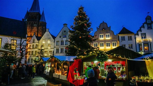Alte Giebelhäuser, ein großer Kirchturm und ein festlich geschmückter Weihnachtsbaum thronen über kleinen Verkaufsständen auf einem Weihnachtsmarkt.