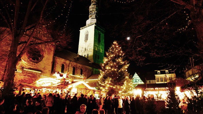 Ein Weihnachtsmarkt.Viele Menschen auf einem Kirchplatz. Helle Lichter. Im Hintergrund ein großer Tannenbaum und eine Kirche.