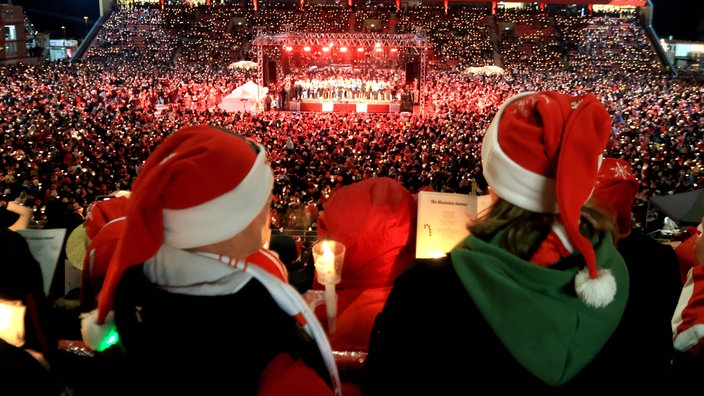 Tausende Menschen stehen mit Liedertexten und Kerzen in den Händen und Weihnachtsmützen auf dem Kopf in einem Stadion