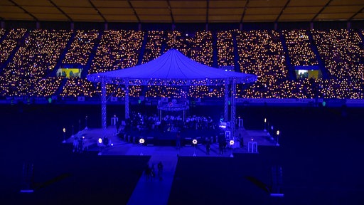 Im Stadion auf dem Tivoli in Aachen steht ein Chor unter einem blau beleuchteten Zelt, im Hintergrund strahlen viele Kerzen auf der Tribüne des Stadions
